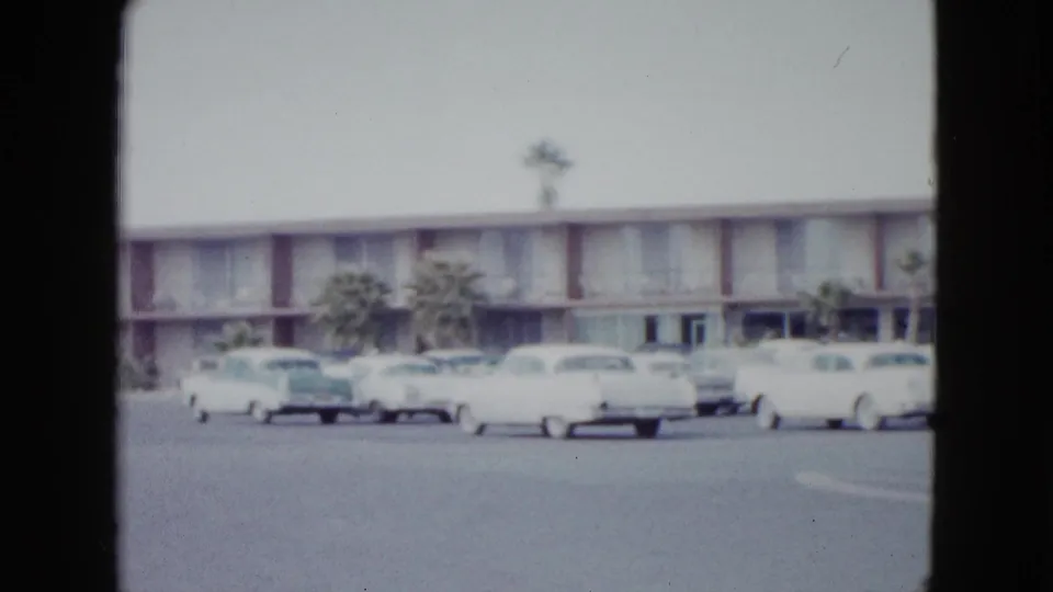 1958 Las Vegas home-movie frame of a motel parking lot with pale 1950s cars and palm trees near the entrance.