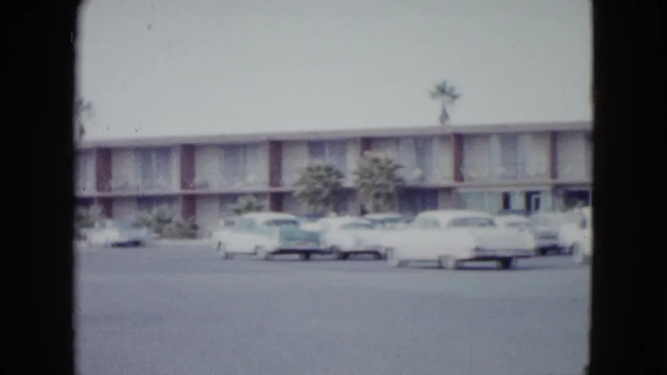 Las Vegas motel exterior in 1958, seen across a parking area with classic cars and palm trees in soft 8mm color.