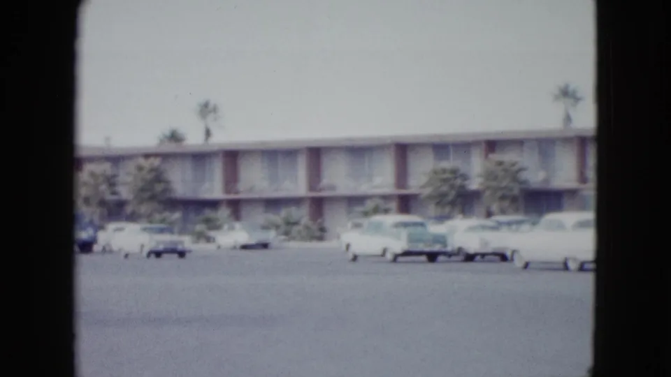 1958 Las Vegas 8mm frame with a motel exterior, palm trees, and parked 1950s cars viewed through dark film borders.