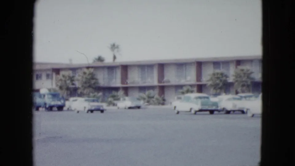 1958 Las Vegas motel parking lot with several parked cars, palm trees, and a low building in soft 8mm color.