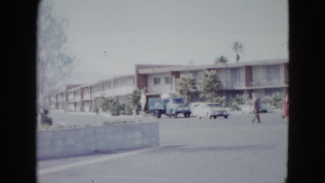 1958 Las Vegas motel parking area with a curb wall, parked vehicles, palm trees, and a pedestrian walking across the lot.