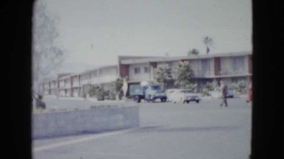 1958 Las Vegas motel parking area with a curb wall, parked vehicles, palm trees, and a pedestrian walking across the lot.