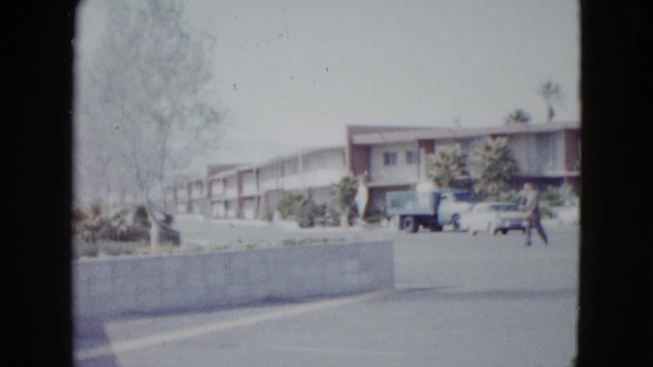 Las Vegas motel parking lot in 1958 with a pedestrian, blue truck, white cars, palms, and a low roadside building.