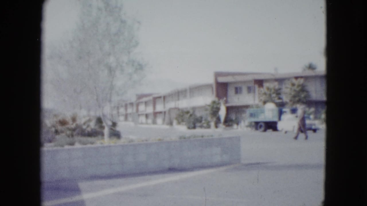 Roadside motel building seen across a drive with a pedestrian and parked cars.