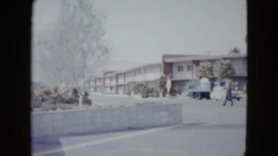 1958 Las Vegas home-movie frame showing a low motel building, driveway, trees, parked cars, and a person walking near the entrance area.