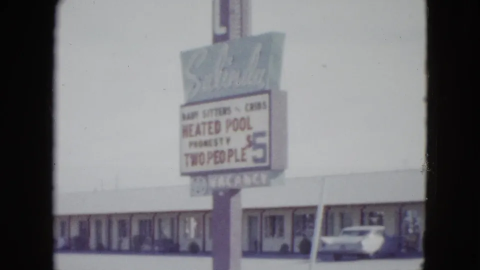 1958 Las Vegas frame with a tall motel sign advertising a heated pool and low room rate, backed by a long single-story motel and parked car.