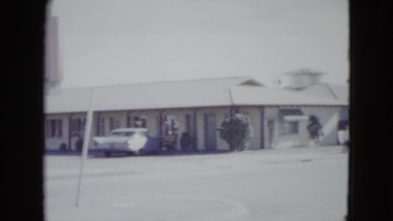 1958 Las Vegas home-movie frame showing a low motel facade, walkway doors, pavement, and a parked white mid-century car.