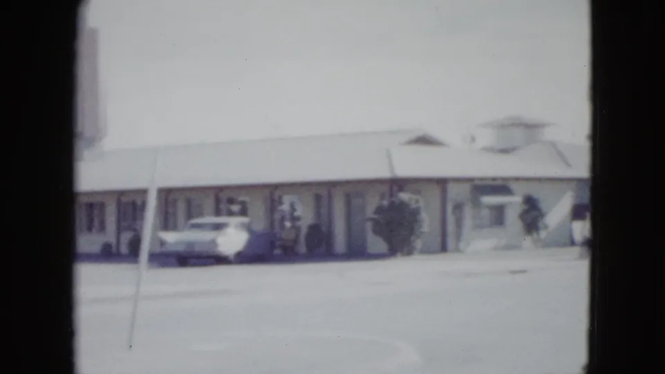 1958 Las Vegas home-movie frame showing a low motel facade, walkway doors, pavement, and a parked white mid-century car.