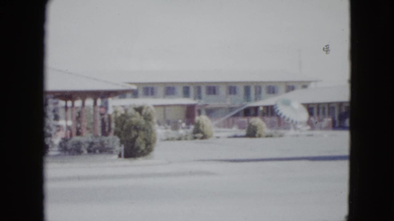 Soft 8mm frame of a low motel facade with shrubs, awnings, and a paved street in front.