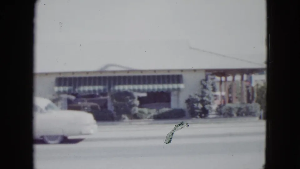 A white 1950s car passes the Las Vegas motel, partially obscuring the striped awning and shrubs.