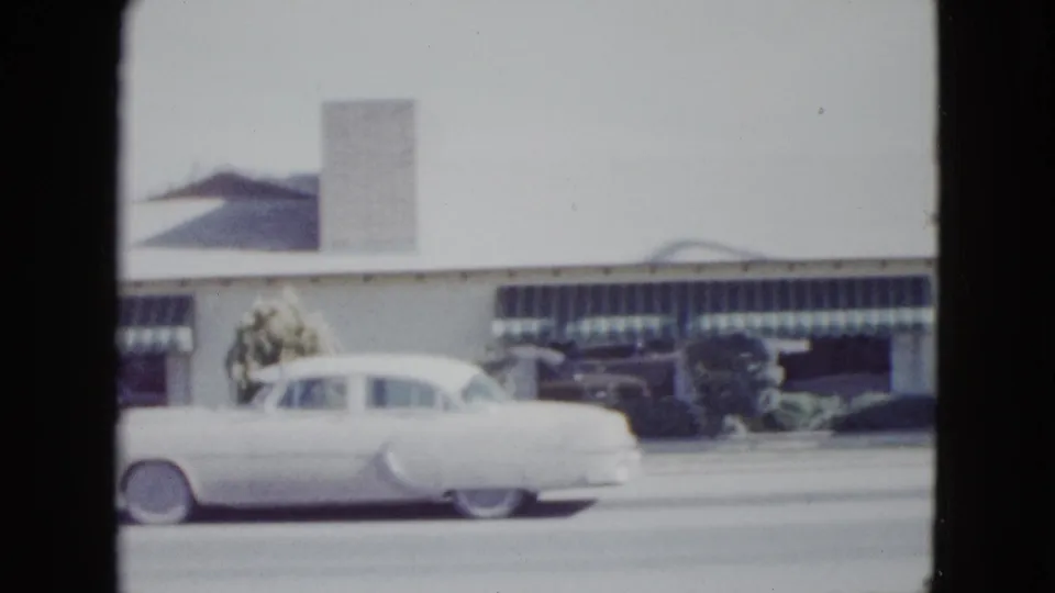 1958 Las Vegas roadside motel view with a white car, striped awnings, shrubs, and a tall sign structure above the roofline.