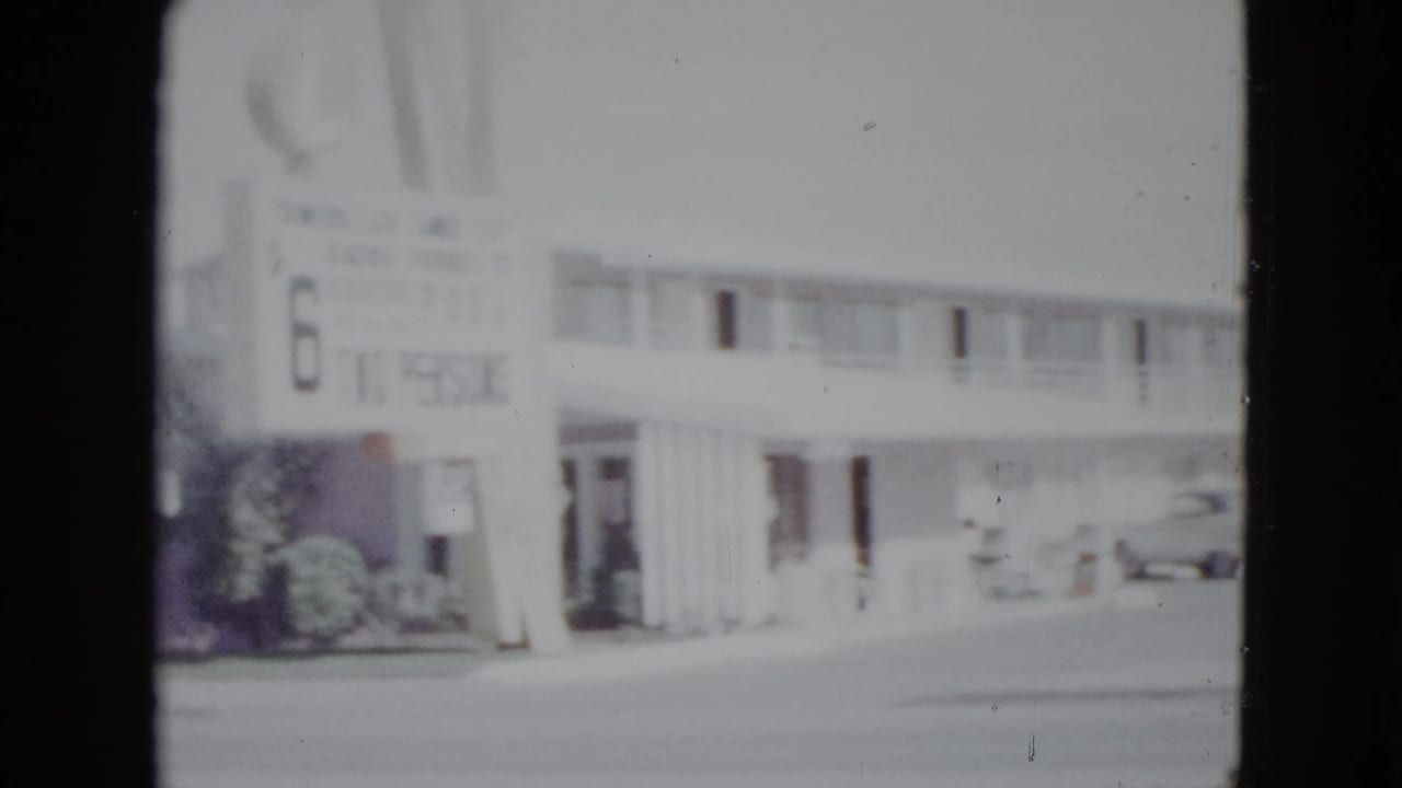 Soft 8mm view of a low-rise motel across a wide street with parked cars and a roadside sign.