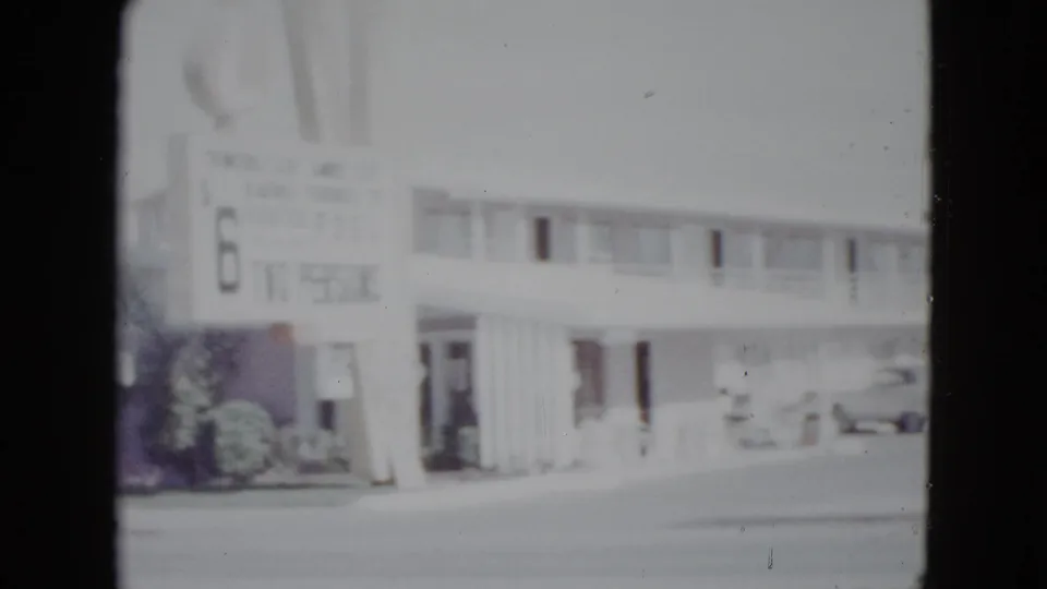 1958 Las Vegas home-movie frame showing a low-rise motel frontage, a sign near the curb, parked cars, and a broad street in bright exposure.