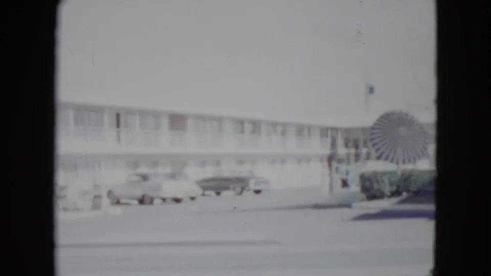 1958 Las Vegas home-movie frame showing a motel across the road, parked cars, shrubbery, and a large circular decorative feature near the building.