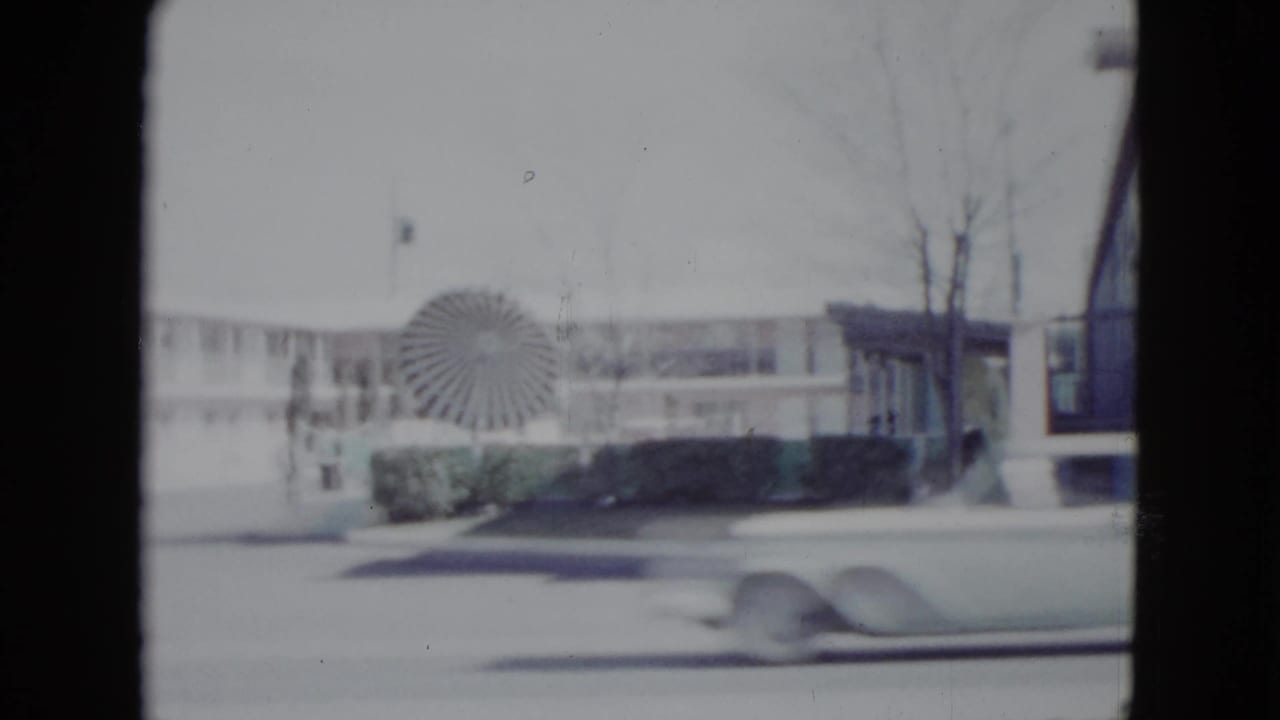 1958 Las Vegas roadside motel scene filmed from a moving vehicle, with a circular sign, low building, bare trees, and a passing car.