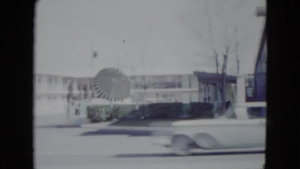 1958 Las Vegas roadside motel scene filmed from a moving vehicle, with a circular sign, low building, bare trees, and a passing car.