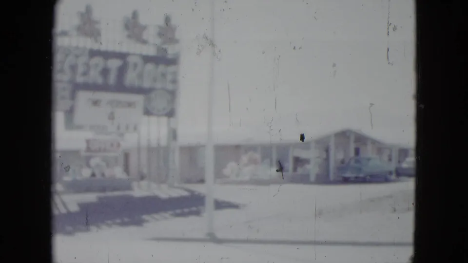 1958 Las Vegas motel exterior with Desert Rose Motel sign, low buildings, curb, and a parked vintage car in faded 8mm color.