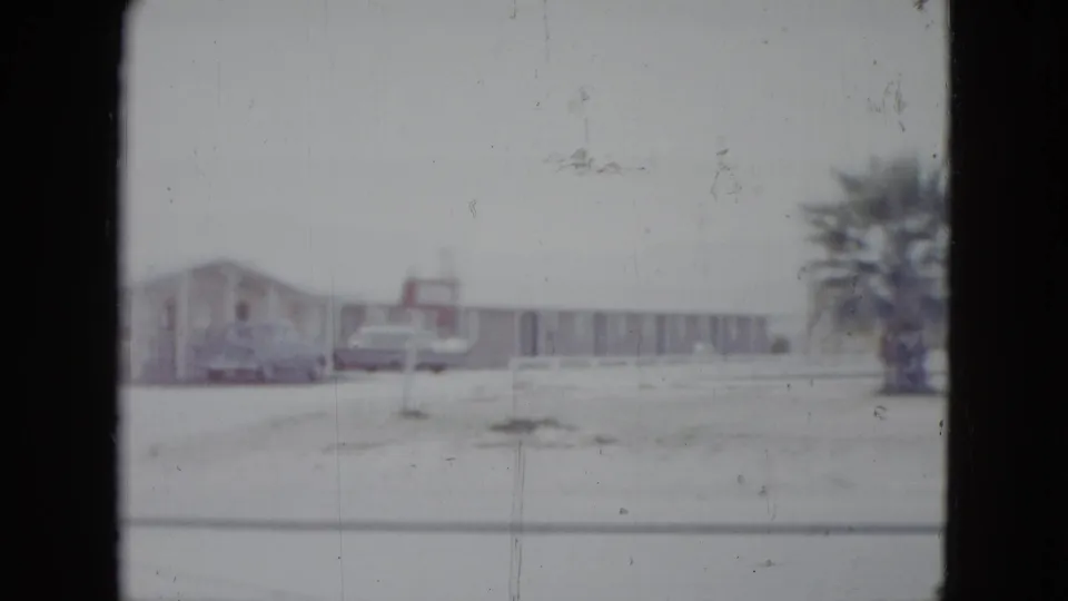 1958 Las Vegas roadside motel view with vintage cars, low lodging buildings, sparse ground, and palm trees in soft 8mm focus.