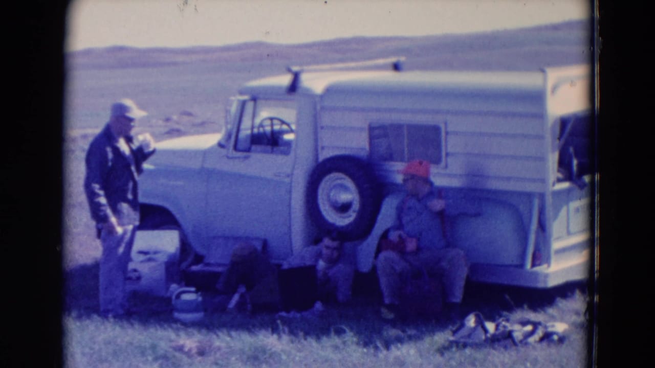 Two adult men pause beside a white truck or camper vehicle in 1958 Omaha road-trip footage, with bags and supplies nearby.
