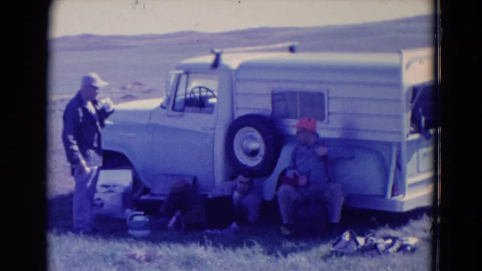 Two adult men pause beside a white truck or camper vehicle in 1958 Omaha road-trip footage, with bags and supplies nearby.