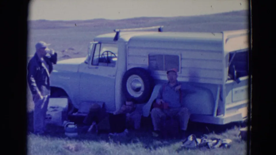 Adult men rest beside a white truck or camper vehicle in 1958 Omaha footage, with a cooler, bags, and kettle-like cookware visible.