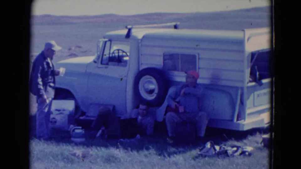 Three adult men appear near a white camper-style truck during a 1958 Omaha road-trip stop, with supplies spread on the grass.