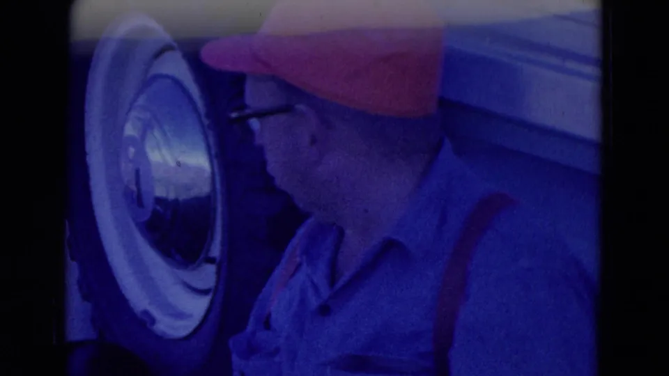1958 Omaha home-movie frame showing a man in a red cap and glasses beside a light-colored truck wheel during tire inspection.