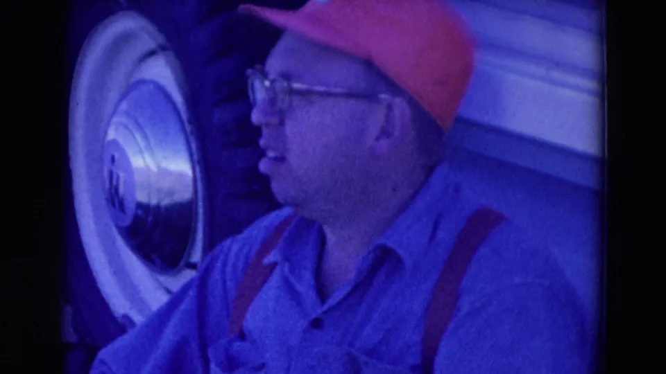 1958 Omaha frame of a man in glasses and red cap beside a whitewall truck tire and light-colored vehicle body.