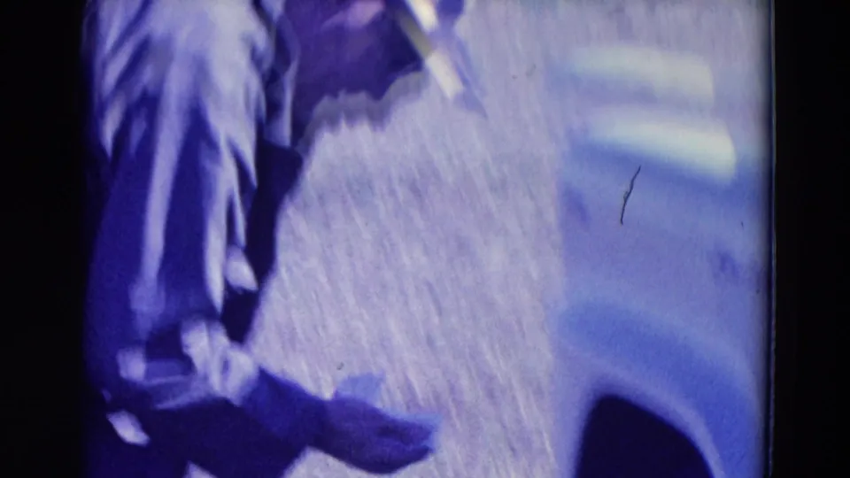 1958 Omaha home-movie frame showing a hunter's lower body near grass and a light-colored vehicle edge, with soft 8mm color shift.