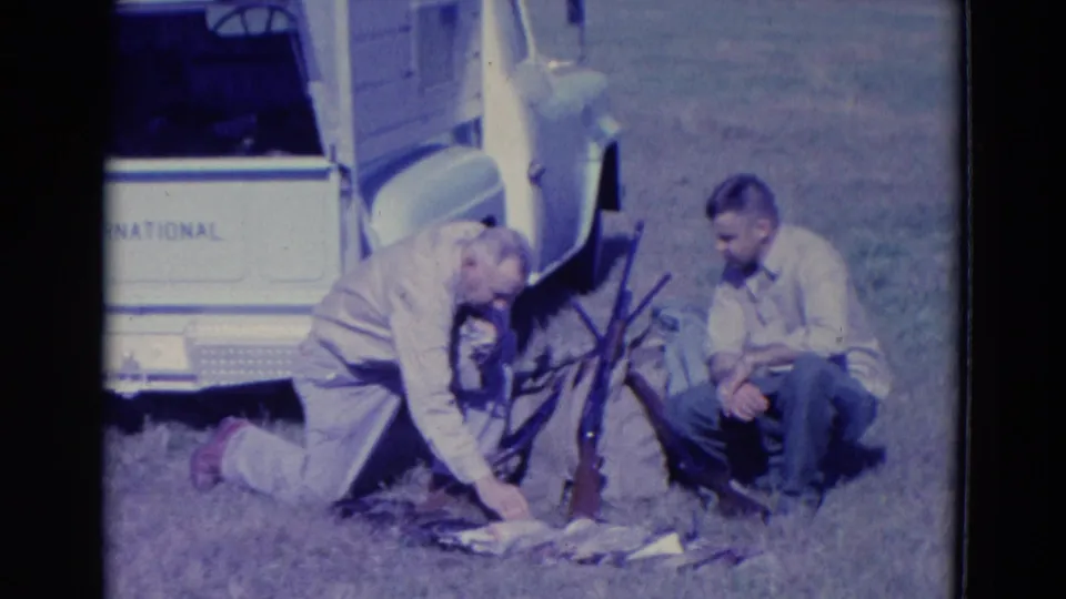 1958 Omaha-area home-movie frame showing two adult hunters beside a white International truck, with rifles upright and harvested ducks on the ground.