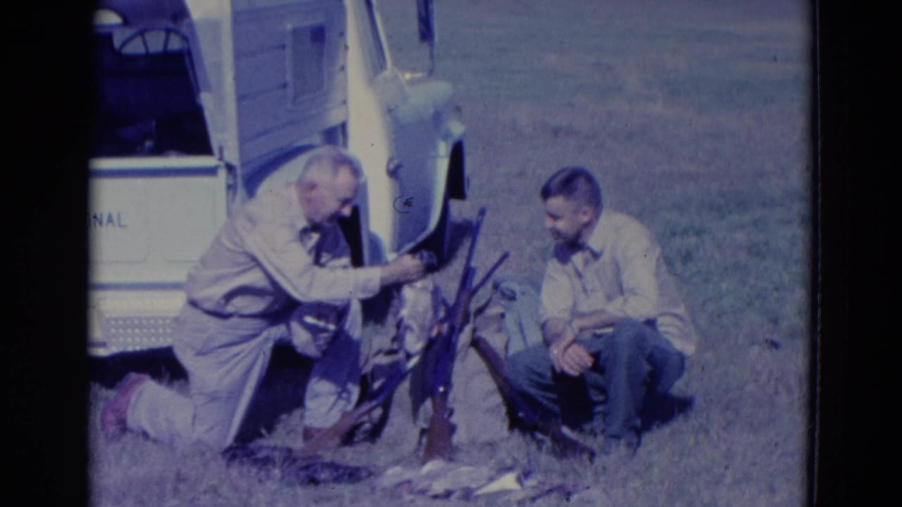 Kneeling hunter reaches toward a duck while another man sits near rifles.
