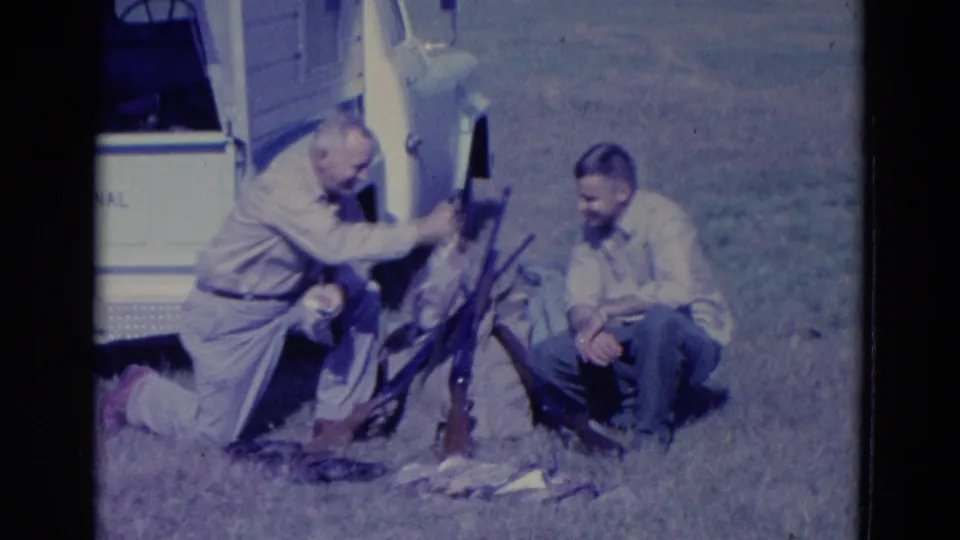 The kneeling hunter raises a duck near several rifles, with a seated man and the white truck visible in the grassy field.