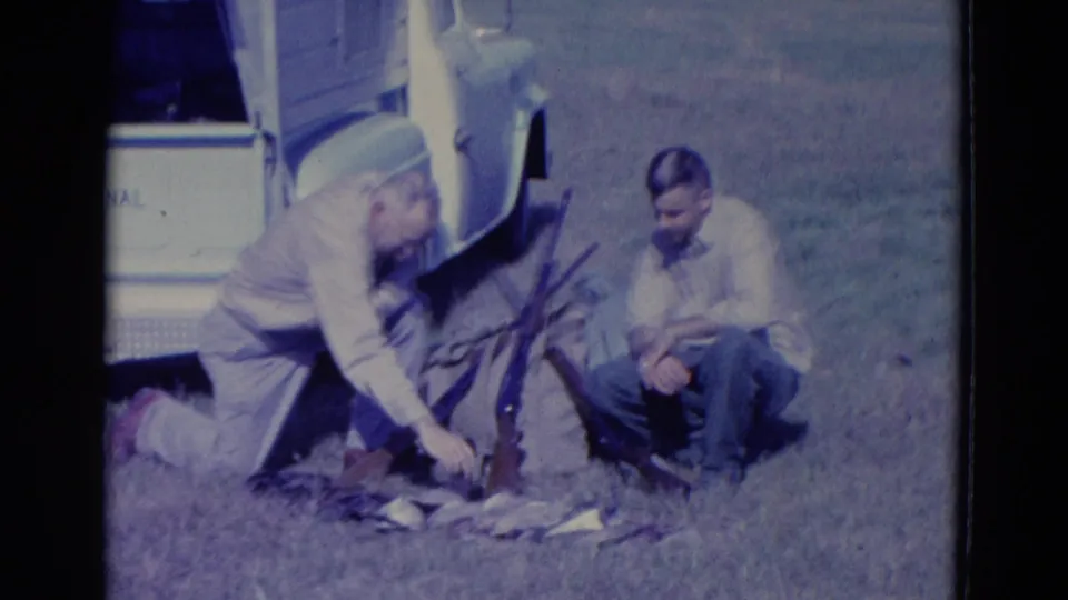 A kneeling hunter reaches down toward the ducks at his feet, with rifles propped nearby and another man seated beside the truck.
