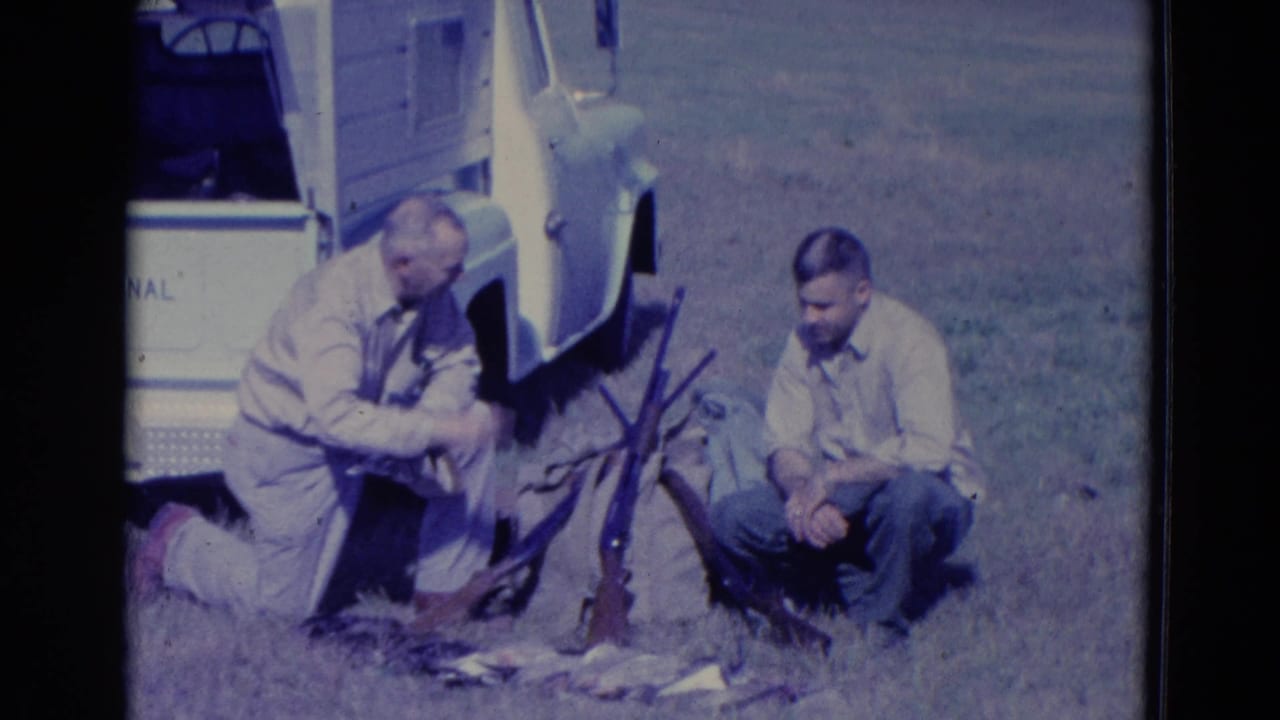 A kneeling man handles a bird while another man sits near shotguns and ducks beside a pickup truck.