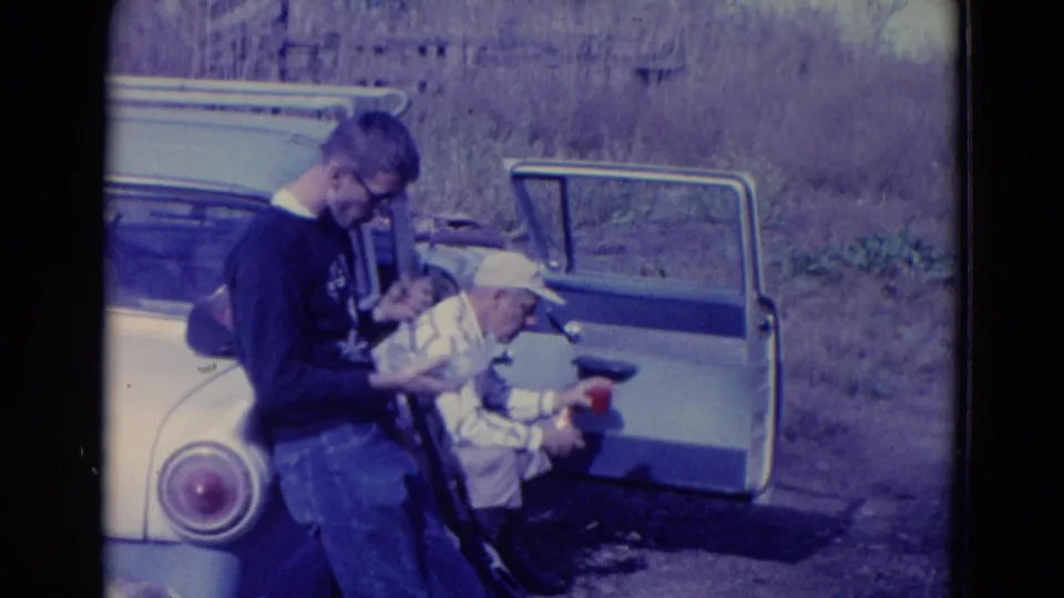 In 1958 Omaha home-movie footage, two adult men stand by a light-colored car, one holding food while the other sits near an open door.