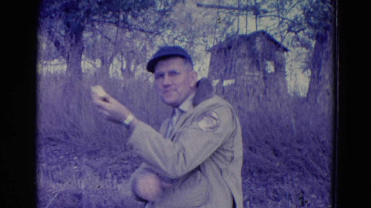 A man in a tan jacket, cap, glasses, and watch faces the camera with a sandwich in a wooded Omaha setting from the 1958 home movie.