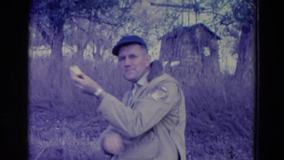 A man in a tan jacket, cap, glasses, and watch faces the camera with a sandwich in a wooded Omaha setting from the 1958 home movie.