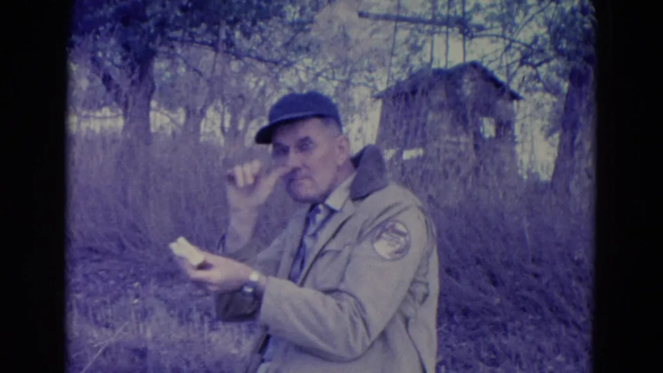 The man in the tan jacket appears to make a playful expression toward the camera while holding a sandwich in the wooded 1958 Omaha scene.