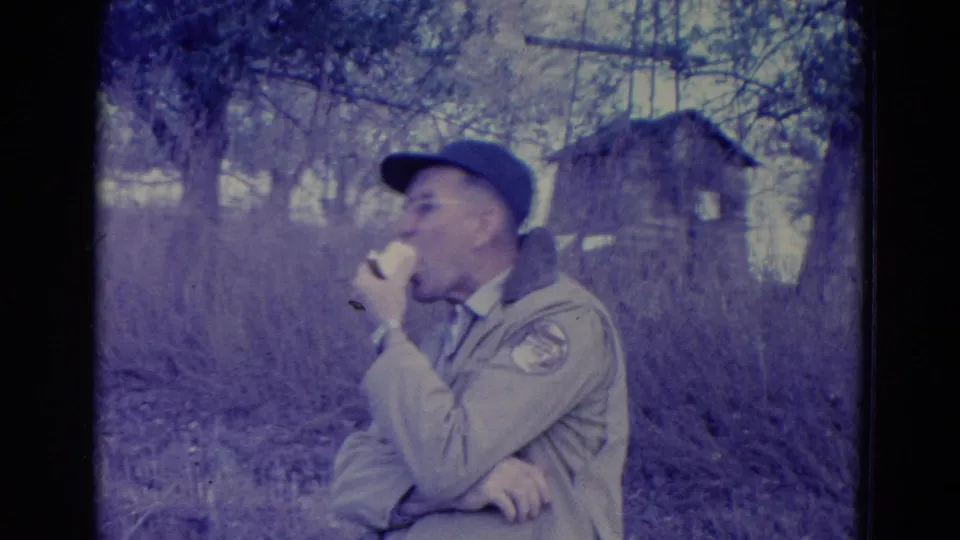 A man in a cap and tan jacket bites into a sandwich, with wooded brush and a small wooden structure behind him in the 1958 Omaha footage.