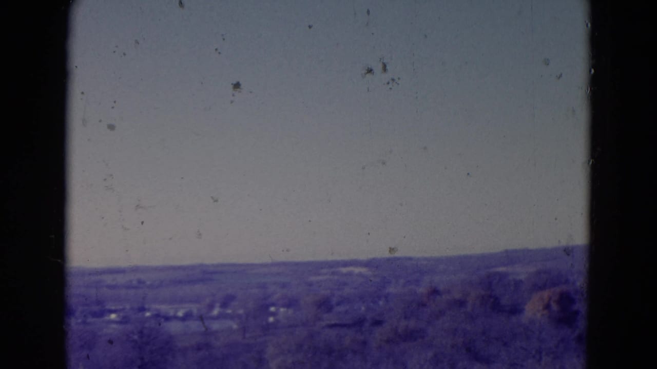 Distant rural hills and tree line seen through dark film or window edges under a hazy sky.