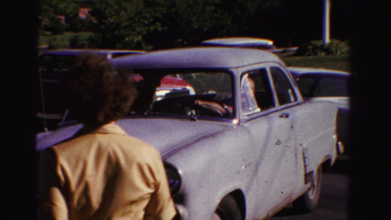 Person with curly hair walks in front of a light-colored sedan in a parking area.