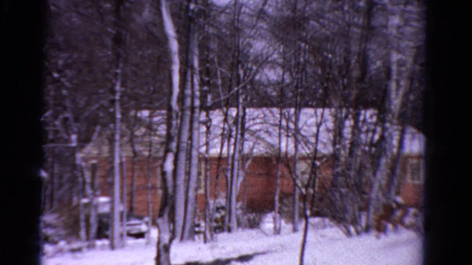 1958 Ontario home-movie frame showing snow-covered buildings, tall bare trees, utility poles, and a partly visible parked car.