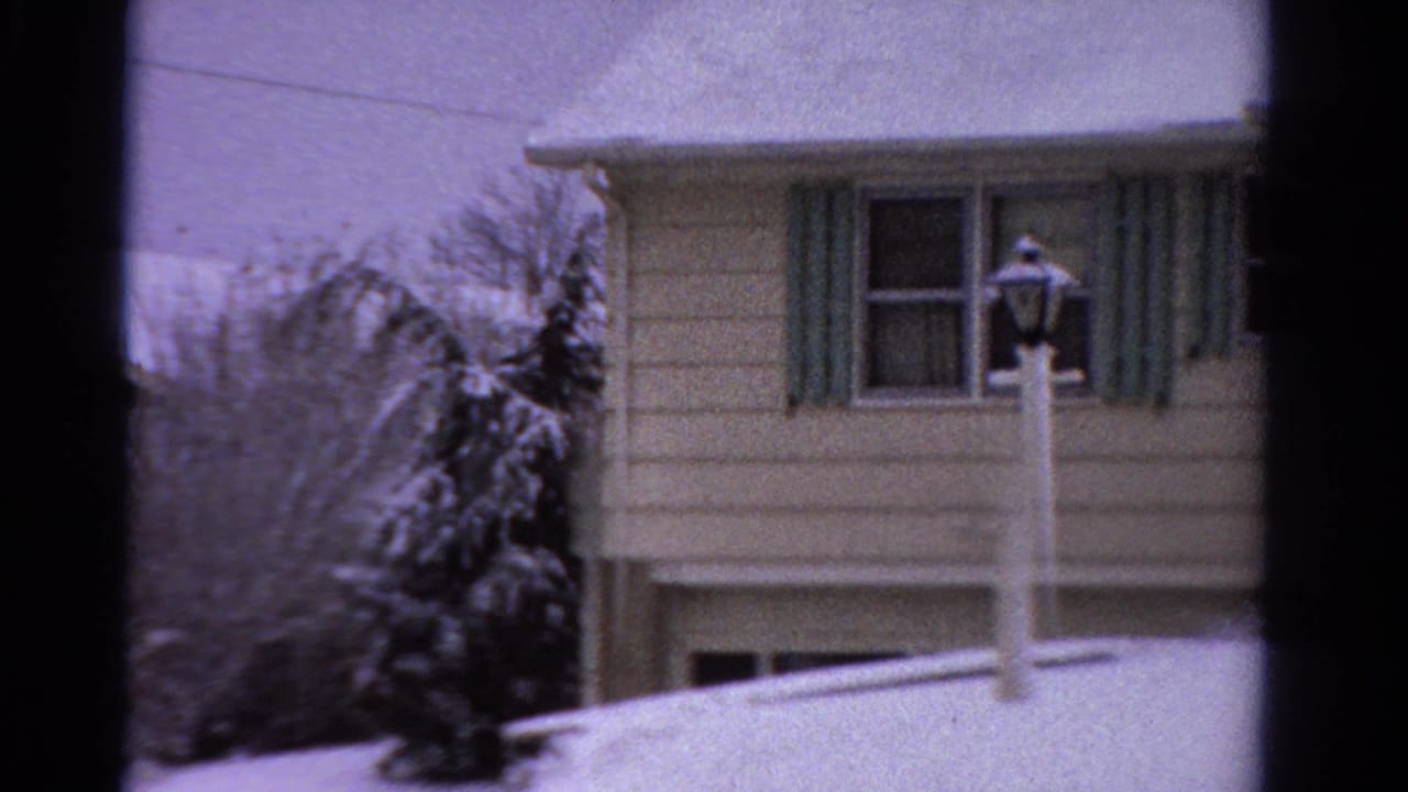 1958 Ontario 8mm frame of a snow-covered clapboard house with green shutters, a front lamppost, shrubs, and dark window edges.