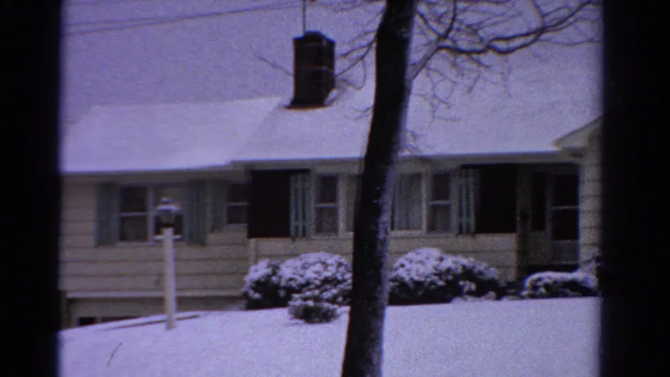 Wider 1958 Ontario 8mm view of the snowy suburban house with a bare tree, chimney, lamppost, bushes, and dark front entry.