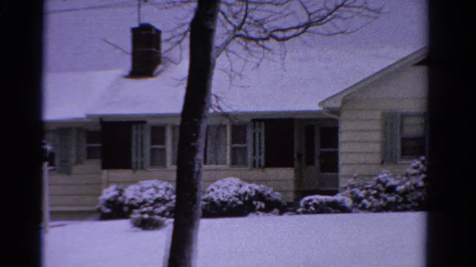 1958 Ontario 8mm frame showing the snowy house from a wider angle with tree trunk, green shutters, bushes, dark entry, and side wall.