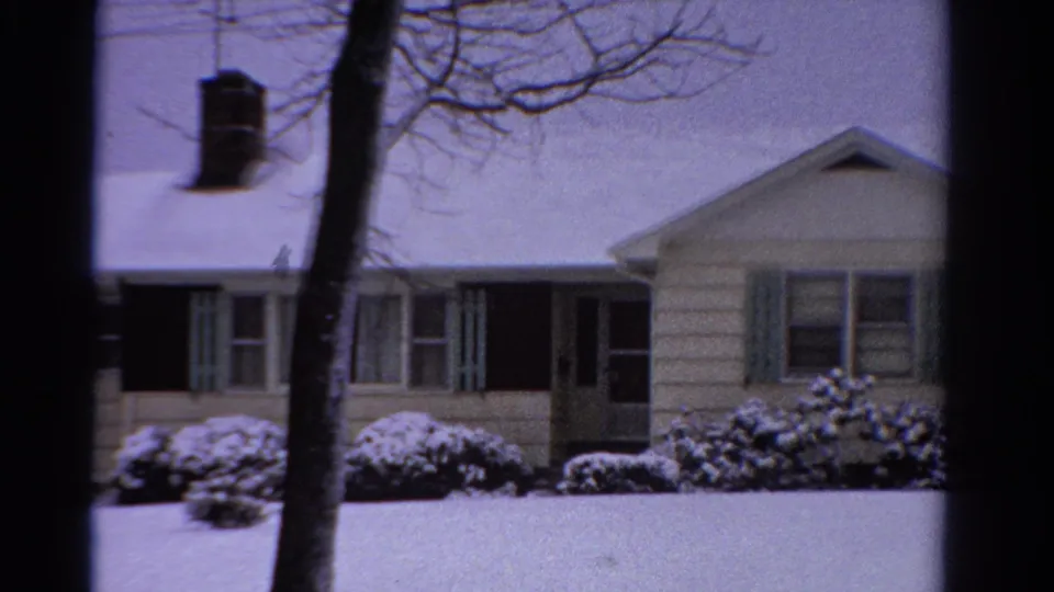 1958 Ontario 8mm frame of a light clapboard house with green shutters, snowy shrubs, and bare tree trunks in front.