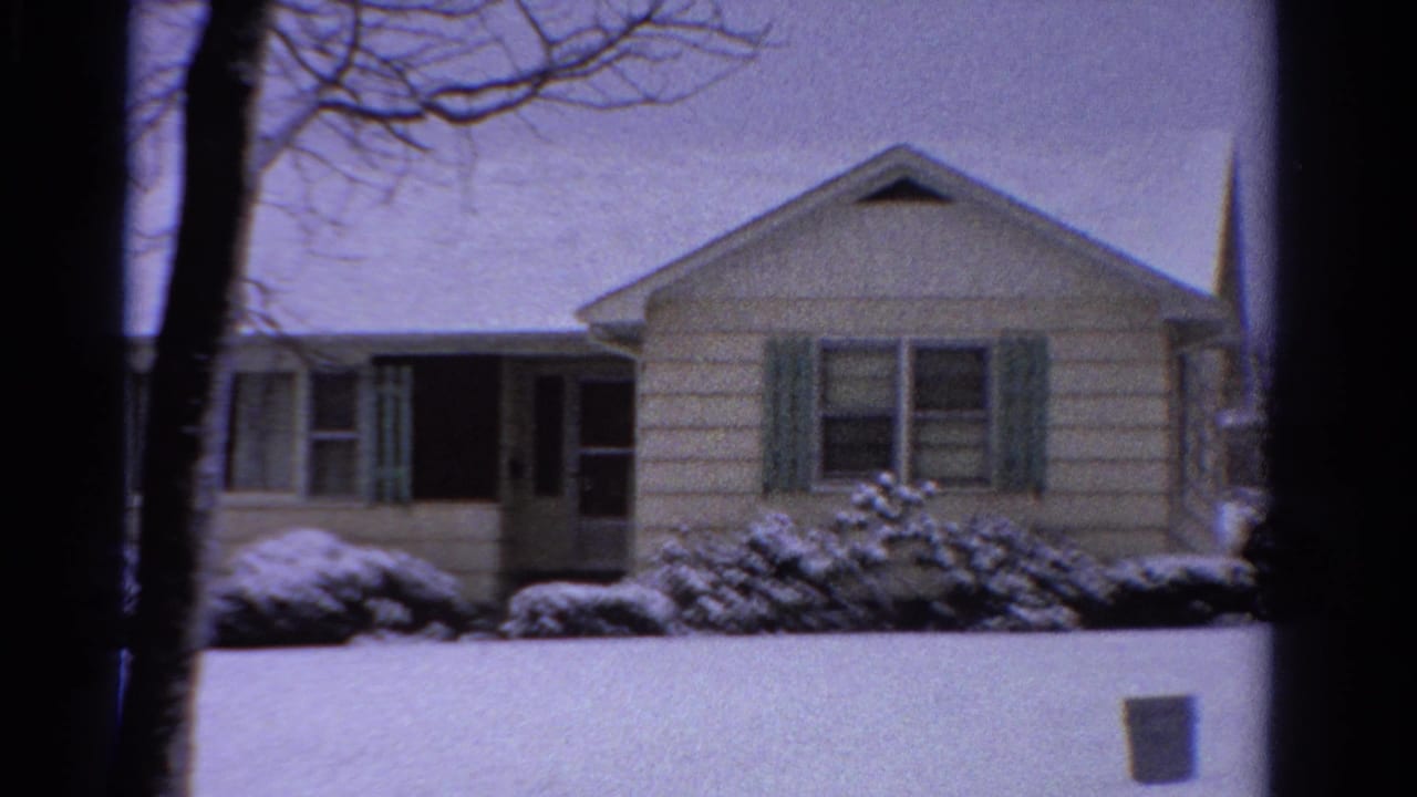 1958 Ontario winter frame showing the front of a clapboard house, snowy bushes, a tree trunk, and a church steeple in the distance.