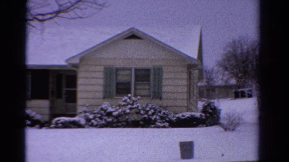 1958 Ontario 8mm frame of a snow-covered house exterior, green shutters, front shrubs, and a partial driveway view.