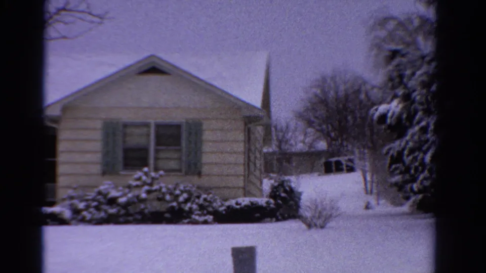 1958 Ontario winter home-movie frame showing a clapboard house, snowy landscaping, driveway, parked vehicle edge, and distant steeple.