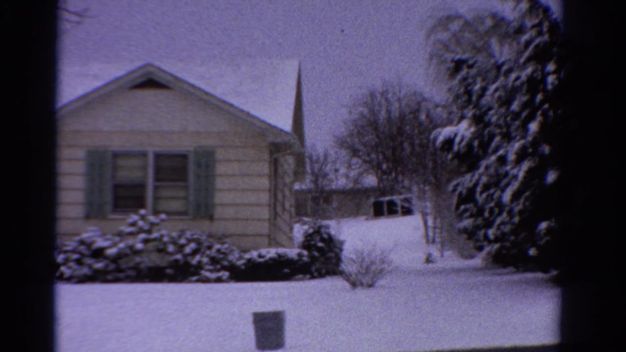 1958 Ontario 8mm frame showing a snow-covered single-story suburban house, green shutters, shrubs, bare trees, and a distant steeple.